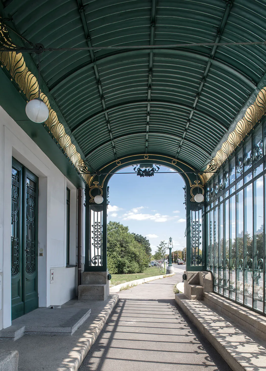 Groen gebogen glazen en metalen dak met gouden decoratieve rand binnen de Otto Wagner Hofpavillon Hietzing, leidend naar een buitenpad.