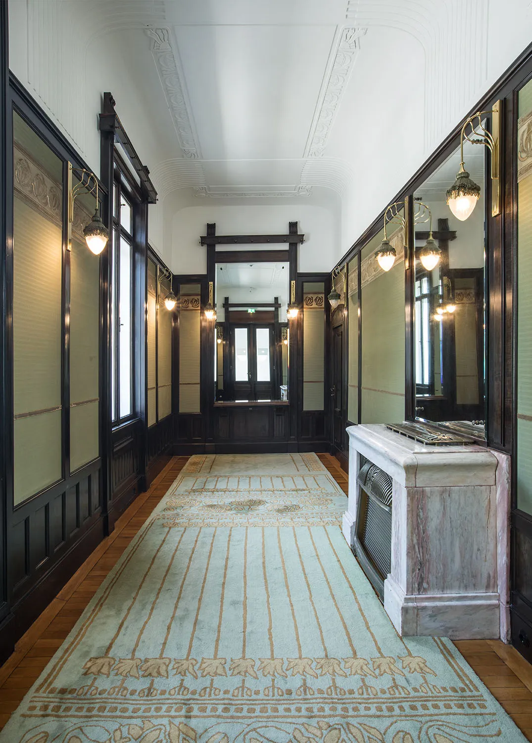 Binnenhal met sierlijk plafond, donkerhouten lambrisering, spiegels en een vloerkleed met patroon