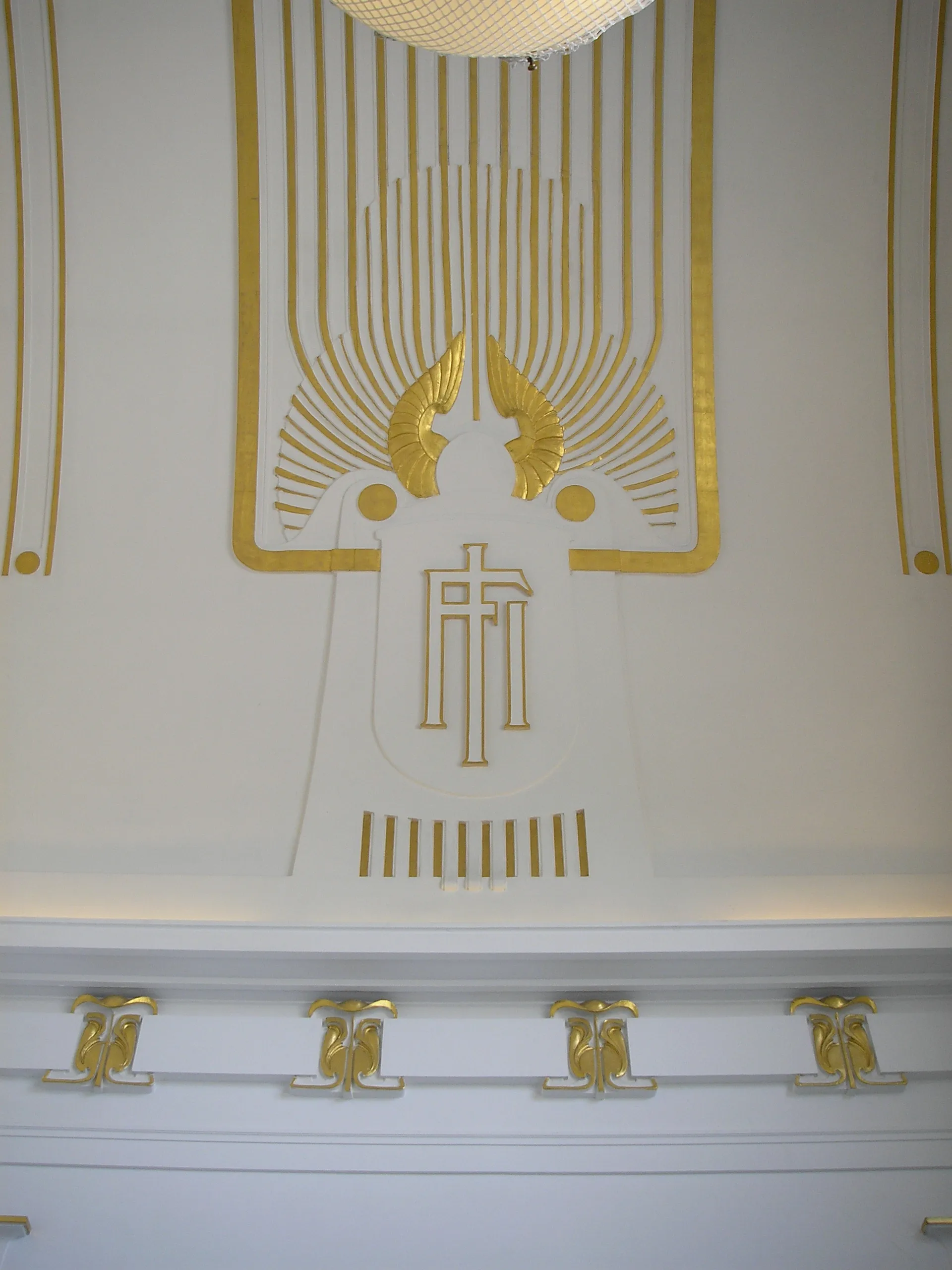 Panel decorativo Art Nouveau dorado con motivos alados y patrones geométricos sobre una pared blanca