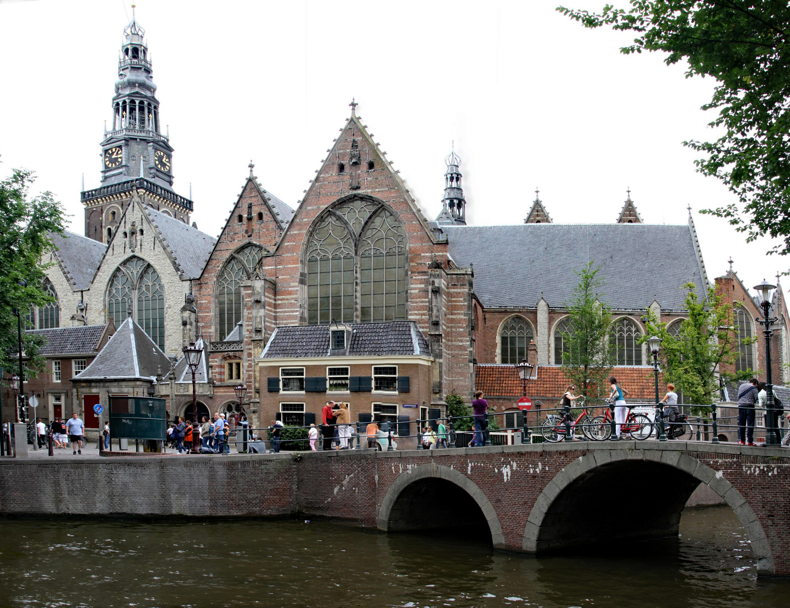 Historic church with a tall spire and Gothic windows, situated beside a canal with a brick bridge and people on the walkway.