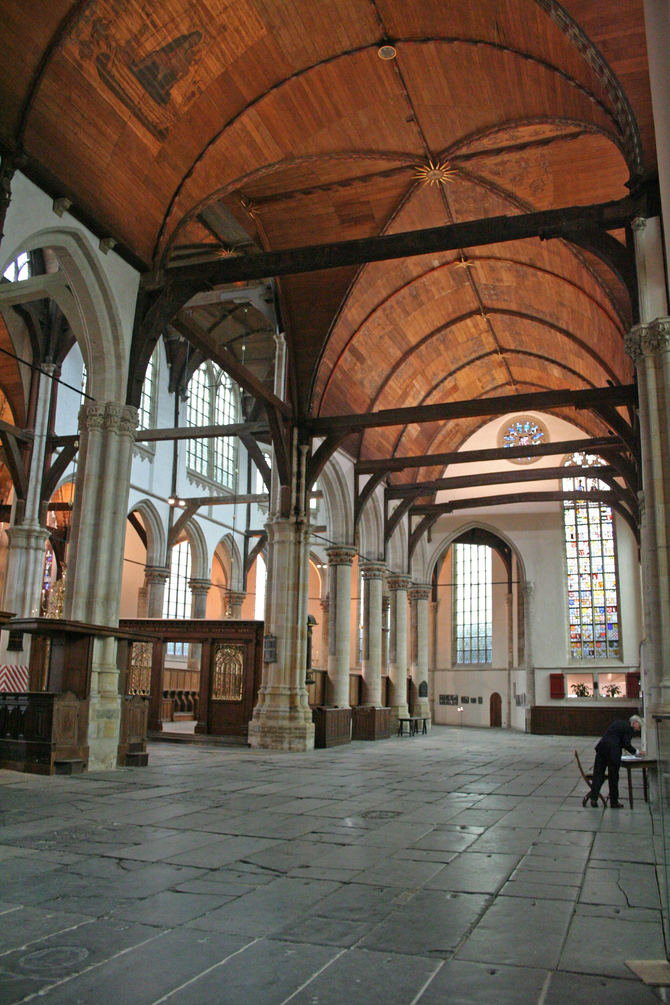Spacious interior of a historic church with high wooden vaulted ceiling, stone pillars, and stained glass windows. One person is visible near the right side.