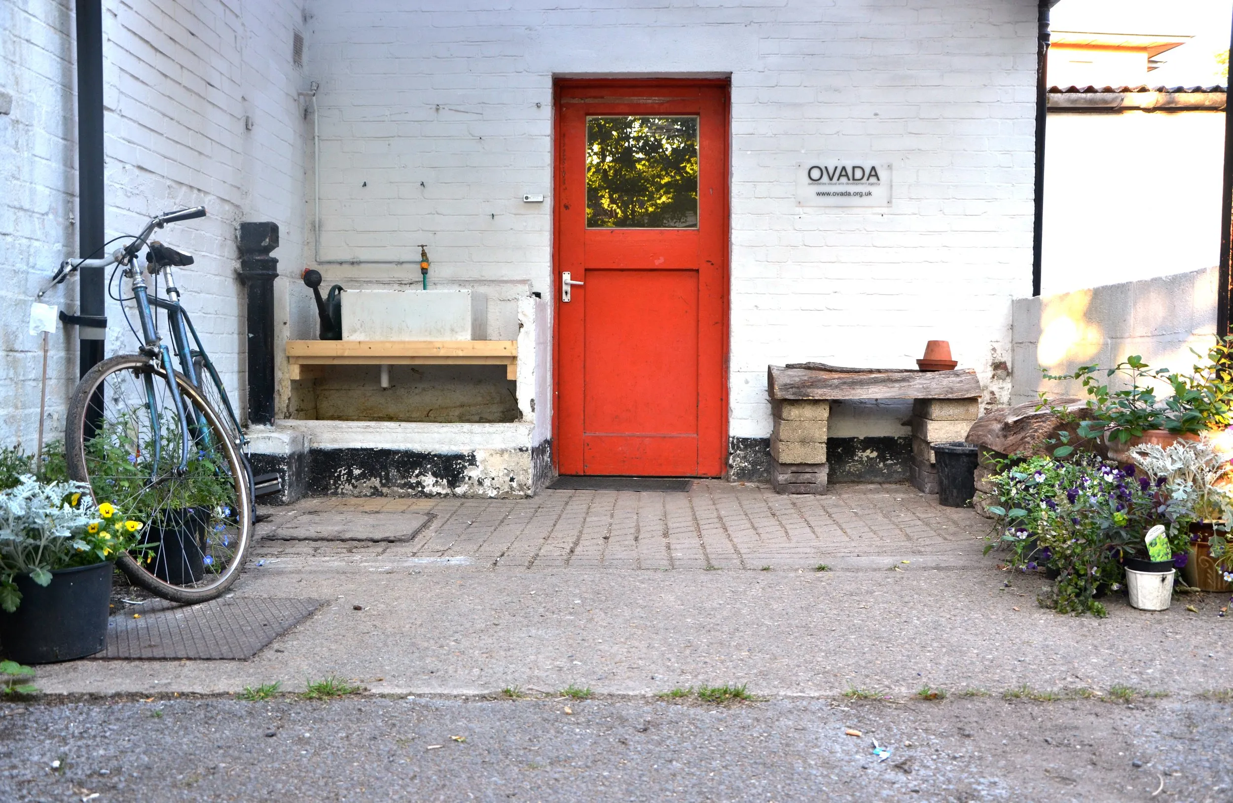 White brick exterior of OVADA Warehouse Gallery with a bright red door, a bicycle leaning against the wall, and potted plants