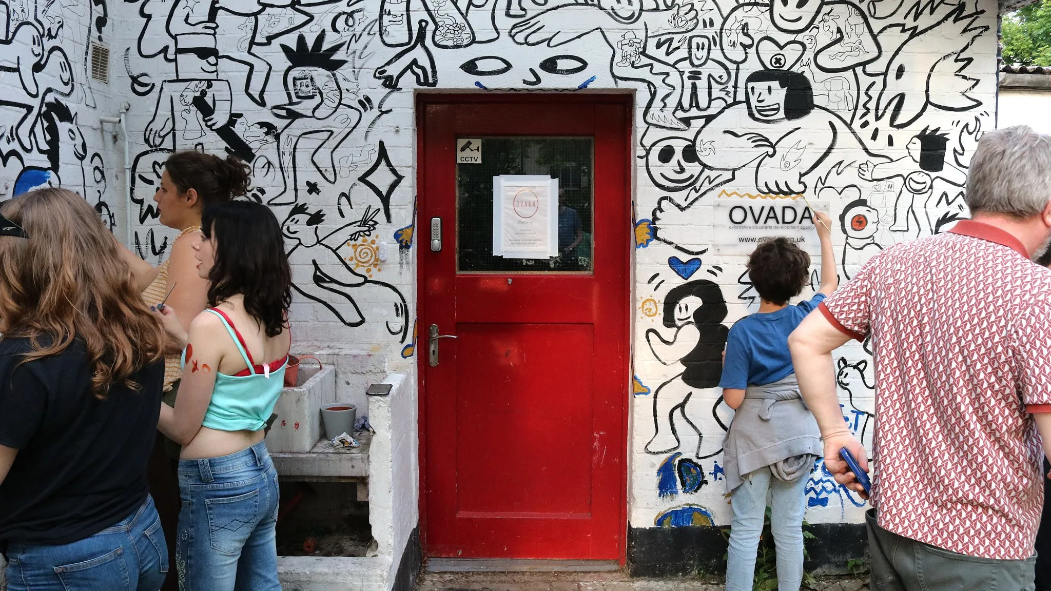 Red door entrance to OVADA Warehouse Gallery surrounded by black-and-white cartoon murals with people viewing the artwork