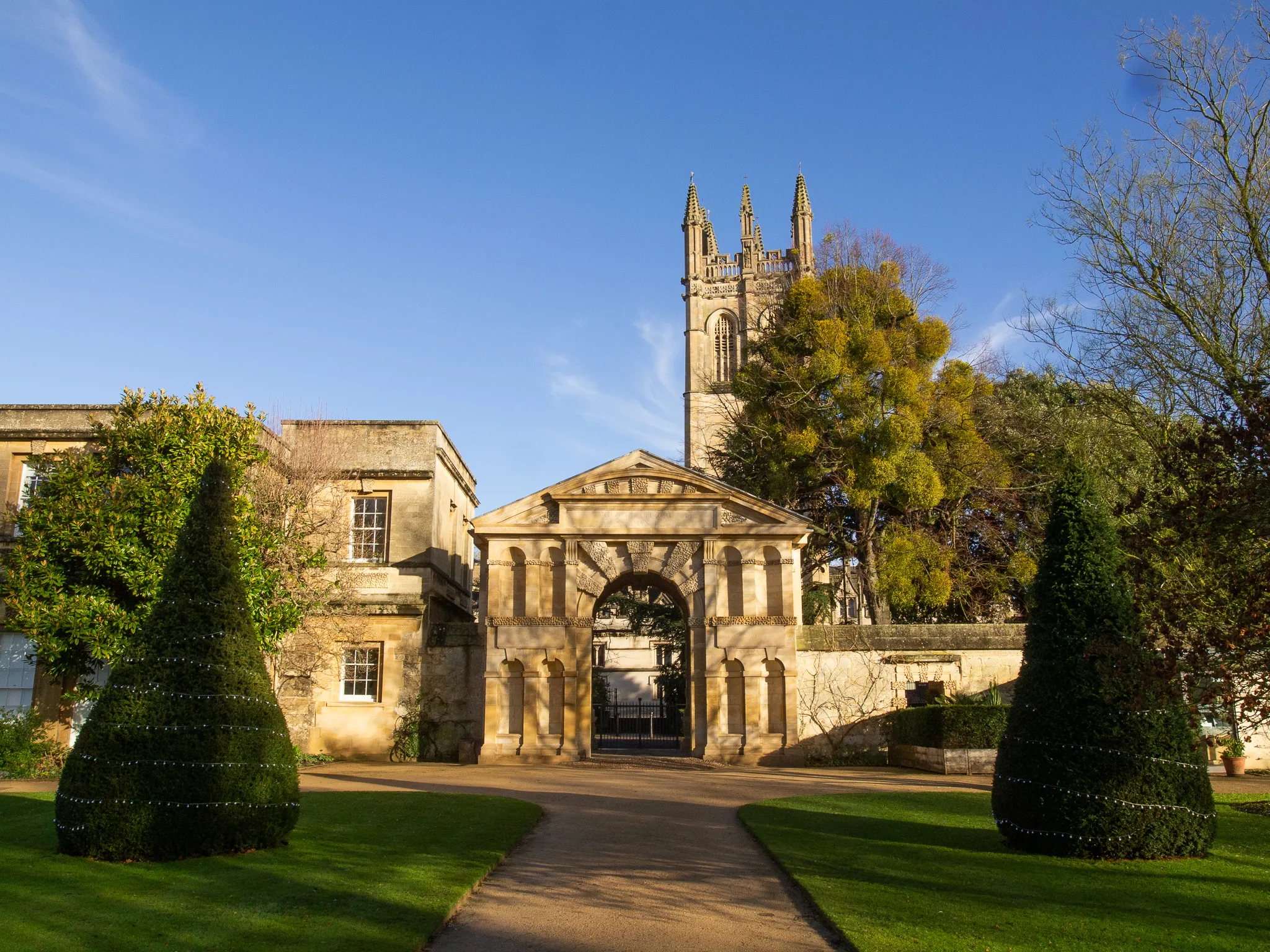 Kamienna brama wejściowa do Oxford Botanic Garden z dwoma stożkowymi cisami flankującymi ścieżkę oraz gotycką wieżą uczelni widoczną w tle pod bezchmurnym niebem
