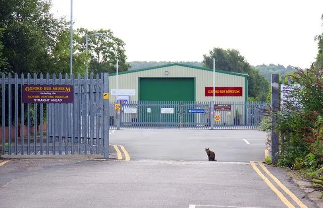 Puerta de entrada al Oxford Bus Museum con un edificio verde al fondo y un gato sentado en el camino de entrada.