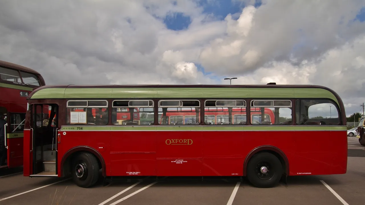 Vista lateral de un autobús antiguo de Oxford de color rojo y verde aparcado al aire libre bajo un cielo nublado, con otro autobús rojo de dos pisos parcialmente visible a la izquierda