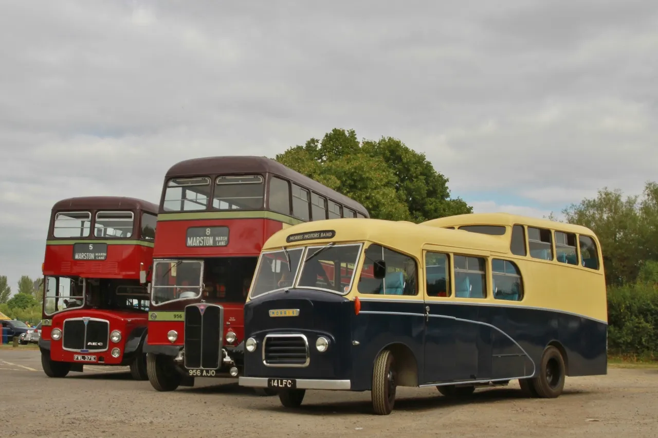 Tres autobuses clásicos aparcados en un terreno de grava bajo un cielo nublado: un autobús de dos pisos rojo, otro de dos pisos rojo y granate, y un autocar Morris FF de una sola planta en color crema y azul.