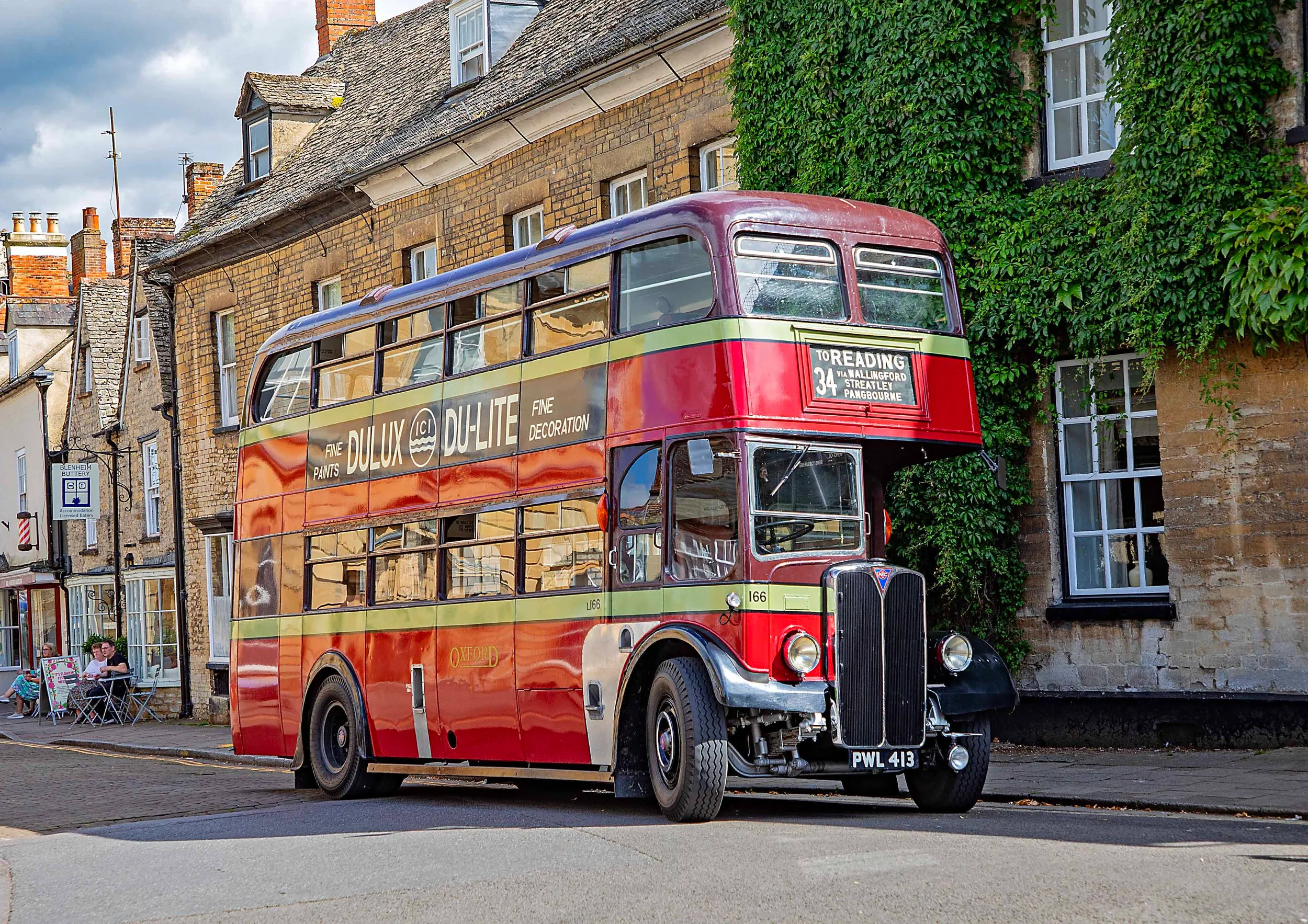 Autobús rojo vintage de dos pisos número 34 a Burford con publicidad de Cadbury's Dualite, aparcado en una calle bordeada de edificios de piedra cubiertos de hiedra
