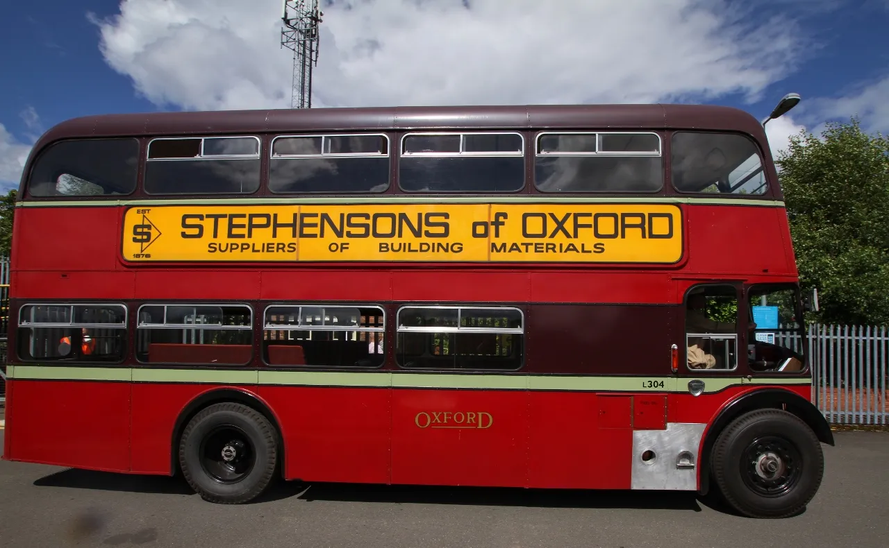 Un autobús clásico de dos pisos de color rojo y granate con un cartel publicitario amarillo de Stephensons of Oxford, estacionado al aire libre bajo un cielo azul con nubes blancas.