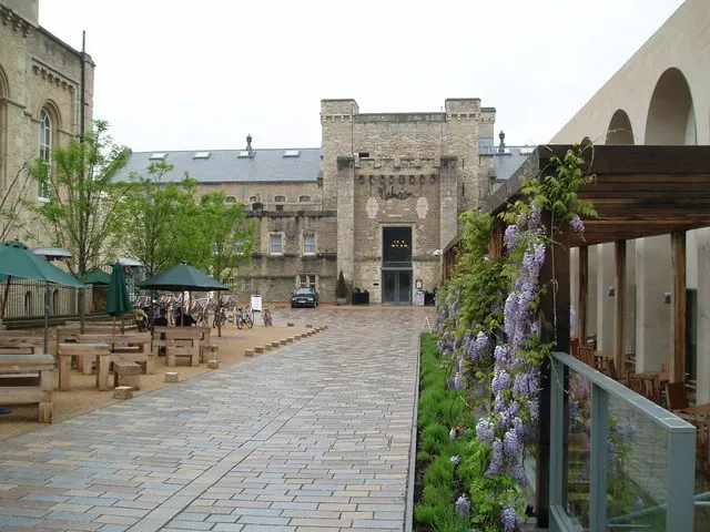 Patio del Oxford Castle & Prison que muestra la fachada del histórico edificio de piedra de la prisión, asientos al aire libre con sombrillas verdes y una pérgola cubierta de glicinias junto a un edificio moderno.