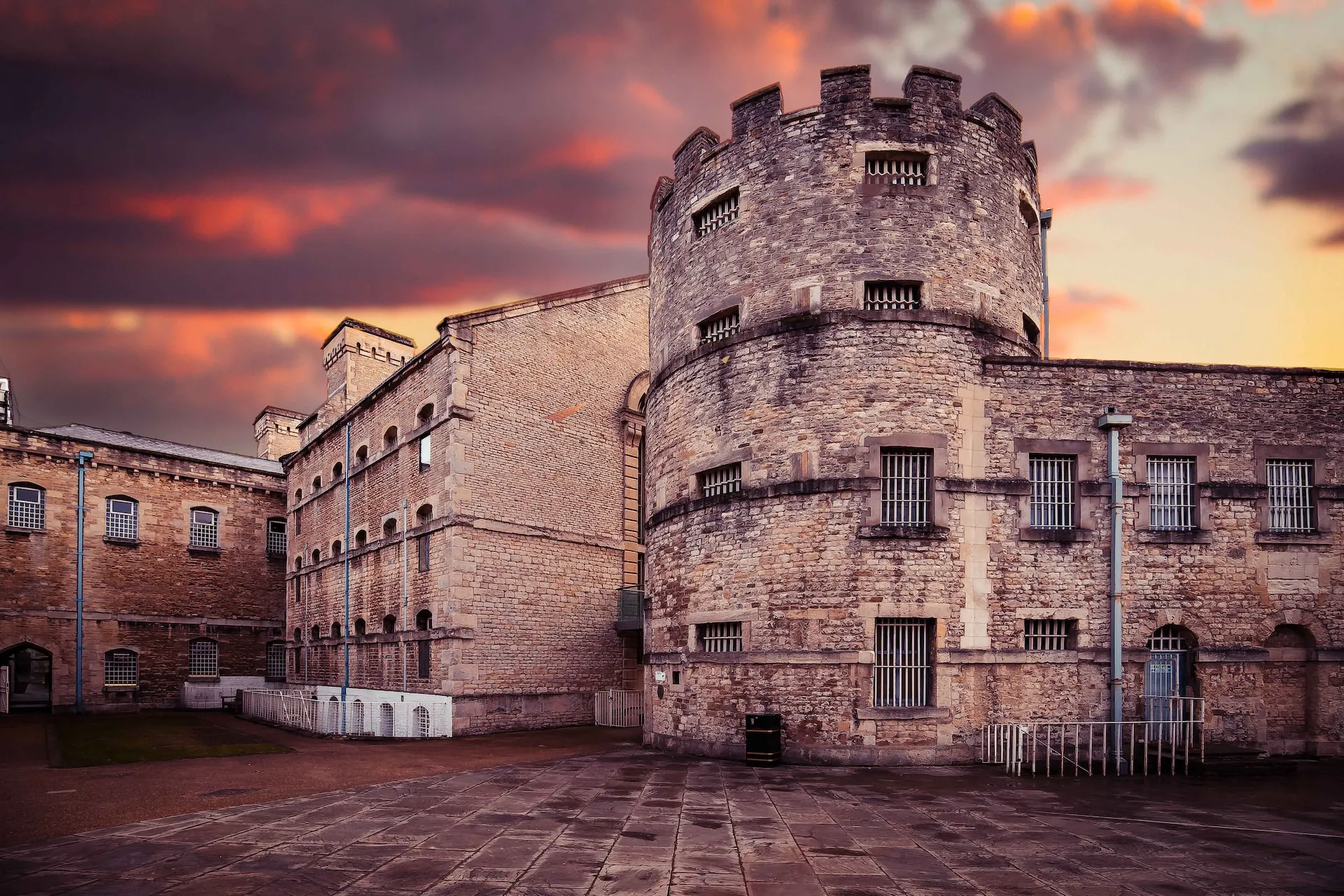 Torre redonda de piedra con almenas y ventanas con rejas, conectada a un edificio de piedra más grande bajo un espectacular cielo de atardecer rosado y naranja, con un patio pavimentado en primer plano