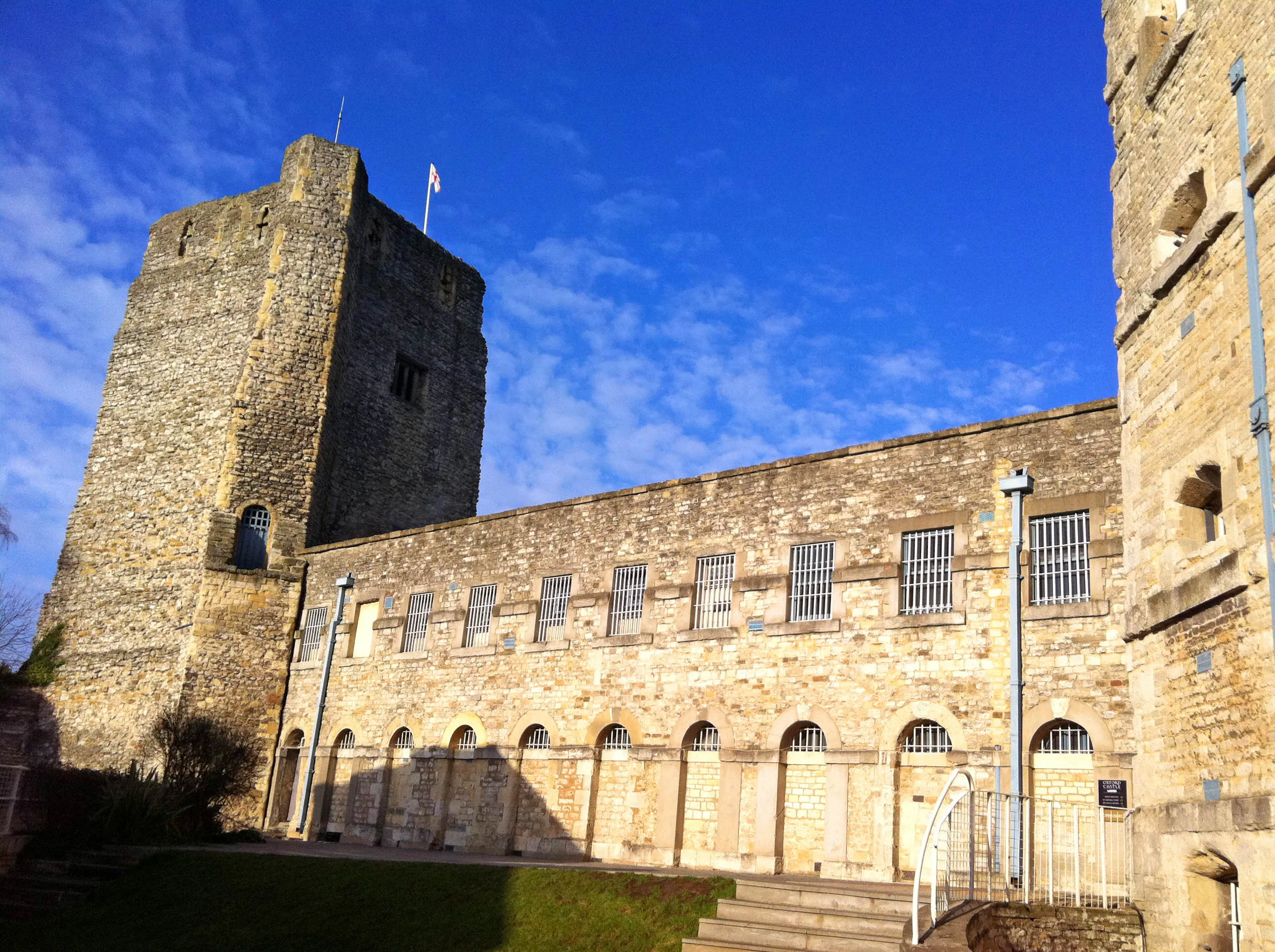 Exterior de piedra del Oxford Castle con una torre cuadrada alta y un edificio largo con ventanas enrejadas bajo un cielo azul.