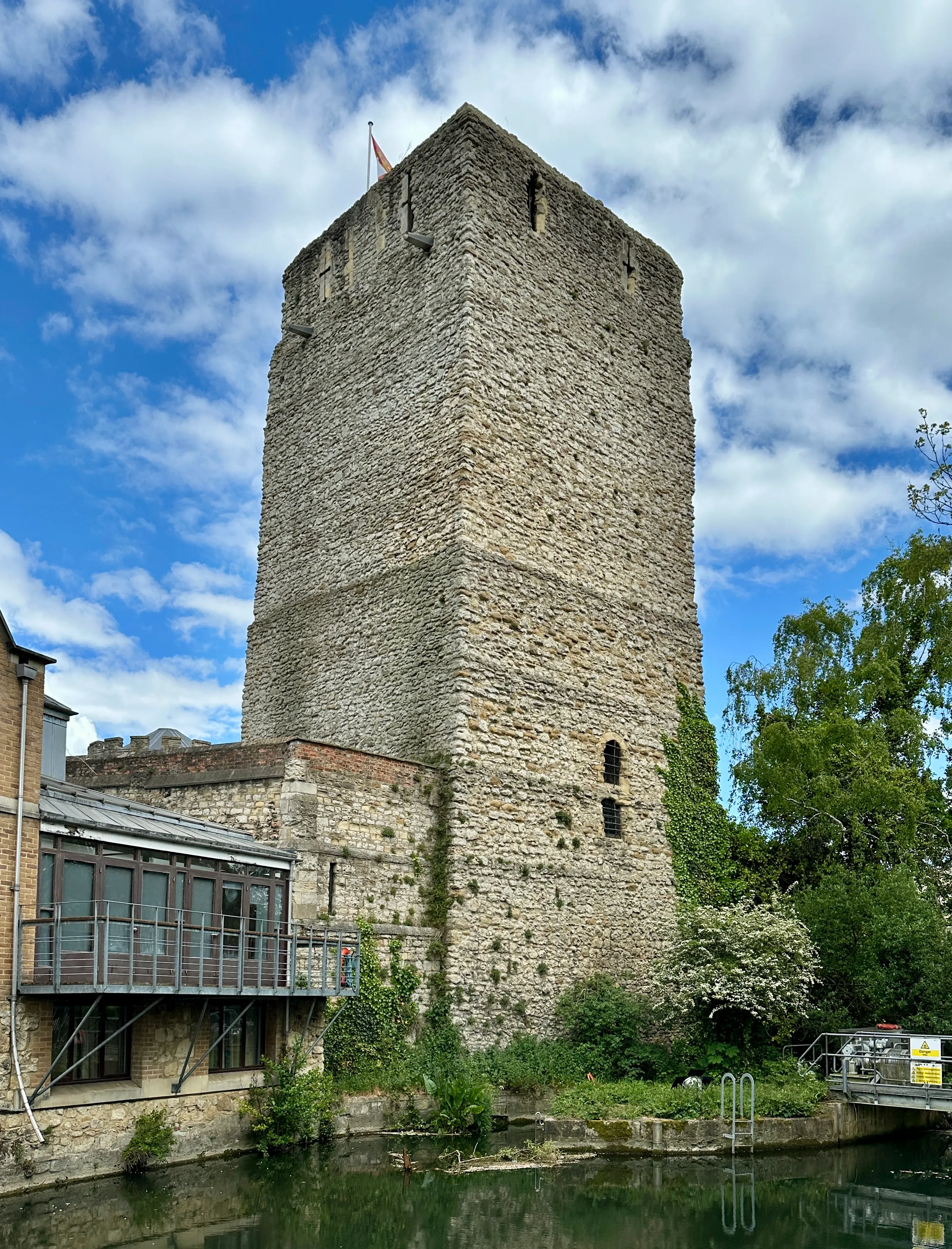 St George's Tower, una alta torre de piedra normanda en el Oxford Castle, que se eleva junto a un canal con una moderna extensión de cristal en su base bajo un cielo azul con nubes