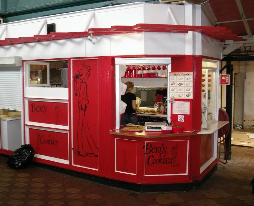 Czerwono-biały kiosk Ben's Cookies z pracownikiem w środku, znajdujący się wewnątrz Oxford Covered Market.