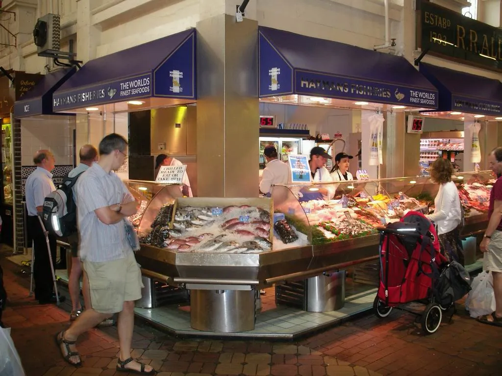 Stoisko rybne z niebieskimi markizami i wystawą świeżych owoców morza wewnątrz Oxford Covered Market, z klientami przeglądającymi ofertę i czerwonym wózkiem dziecięcym w pobliżu