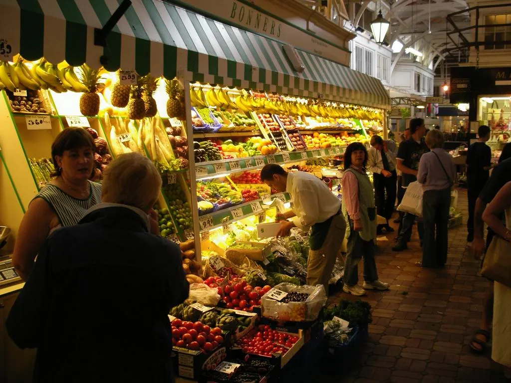 Stoisko z owocami i warzywami wewnątrz Oxford Covered Market, gdzie klienci przeglądają świeże produkty pod zielono-białymi markizami