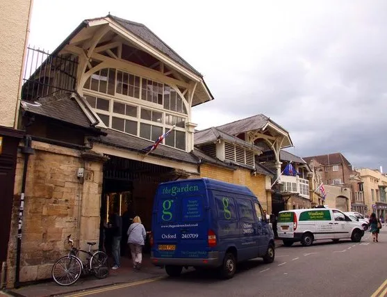 Widok z ulicy na wejście do Oxford Covered Market z charakterystycznym zadaszeniem z drewna i szkła, niebieskim samochodem dostawczym oraz pieszymi.