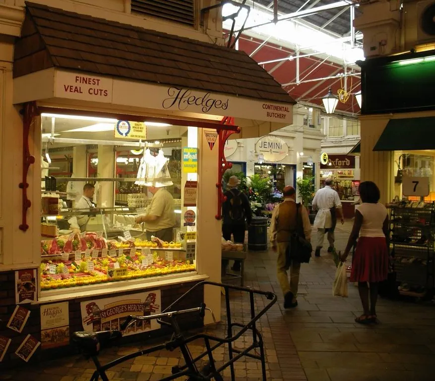 Widok wnętrza Oxford Covered Market z rzeźnią Hodges, szklaną ladą z mięsem oraz kupującymi przechadzającymi się po wyłożonej kafelkami alejce.
