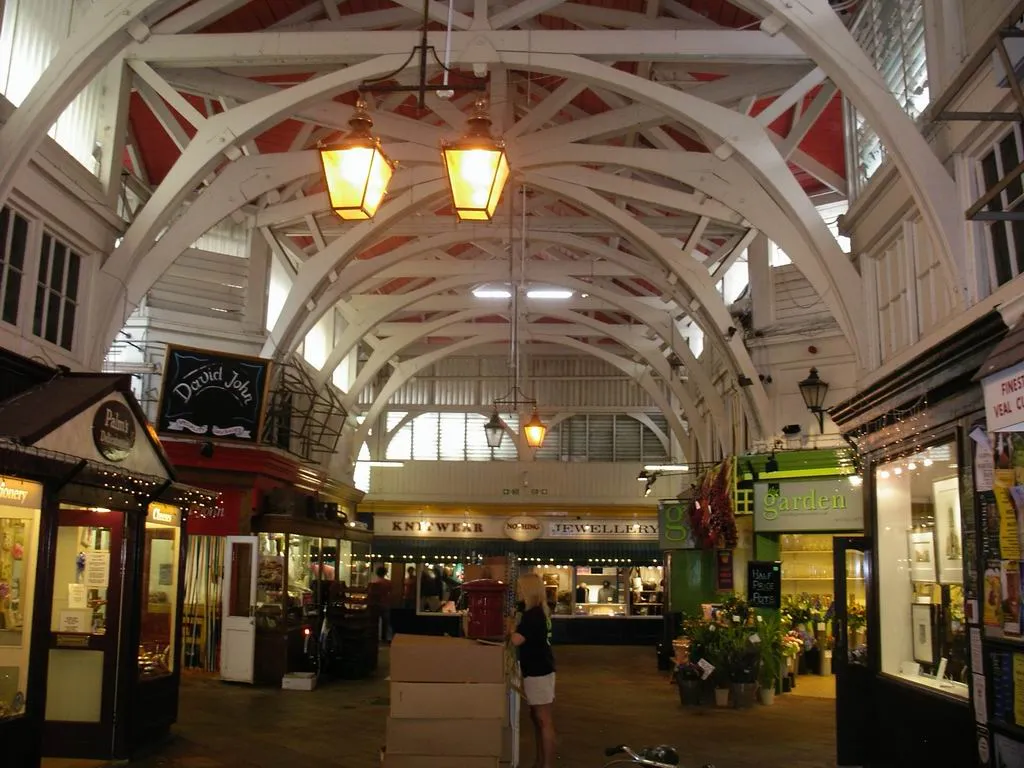 Widok wnętrza Oxford Covered Market przedstawiający łukowate białe belki stropowe z czerwonymi panelami, wiszące latarnie oraz stragany ustawione po obu stronach przejścia