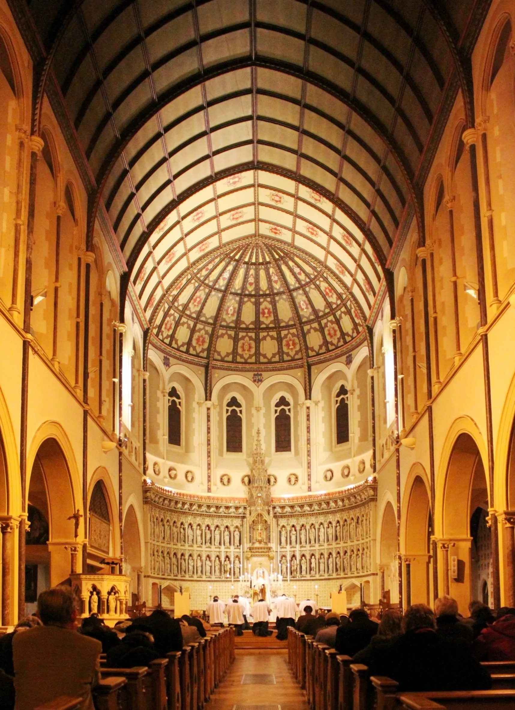 Vista interior del Oxford Oratory que muestra la nave con techo de bóveda de cañón, cúpula decorada sobre el ábside y la congregación sentada en bancos de madera durante un servicio religioso.