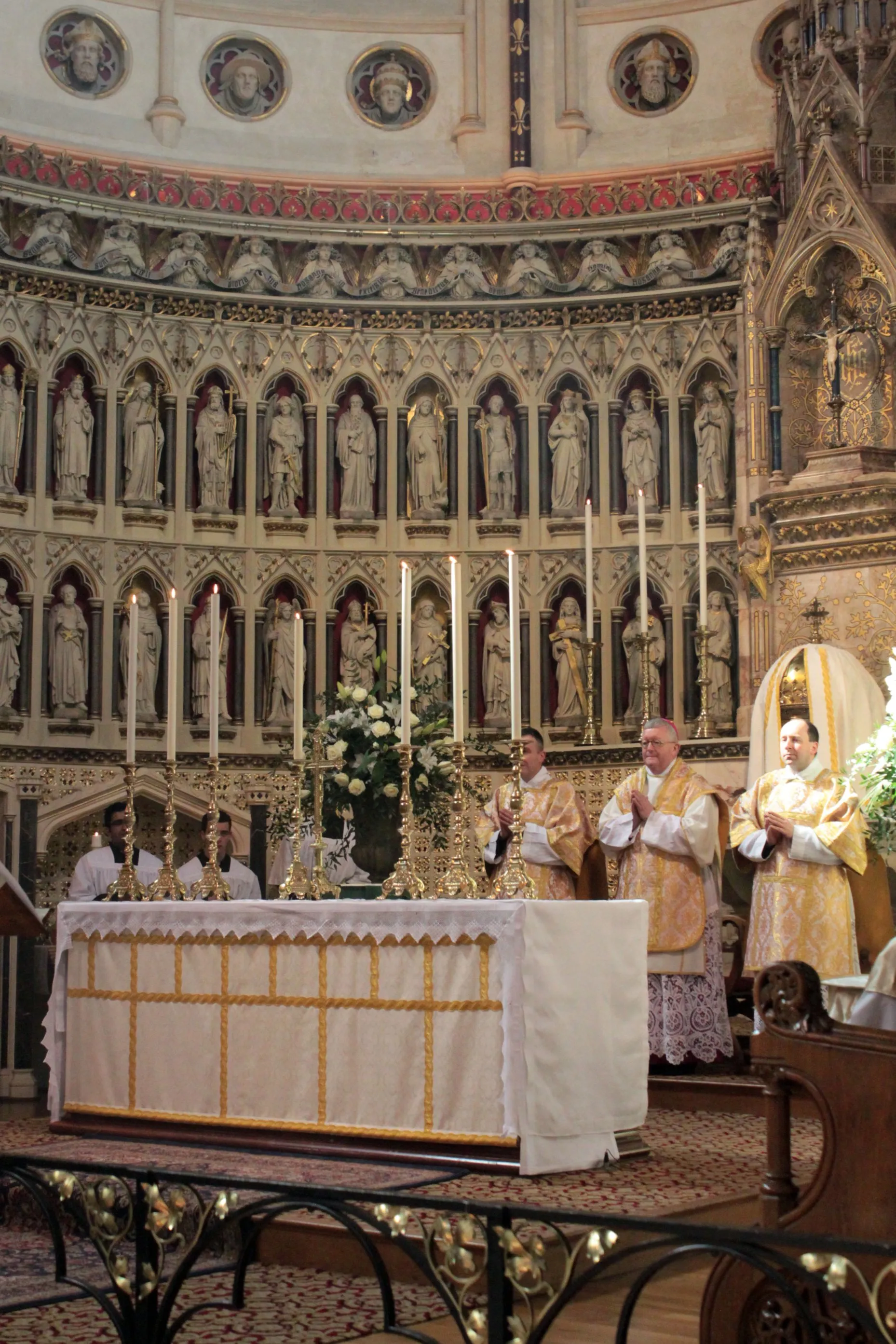 Interior del Oxford Oratory que muestra a los sacerdotes con vestiduras doradas ante un altar iluminado por velas, con un ábside neogótico que presenta filas de estatuas en hornacinas y medallones pintados en la parte superior.