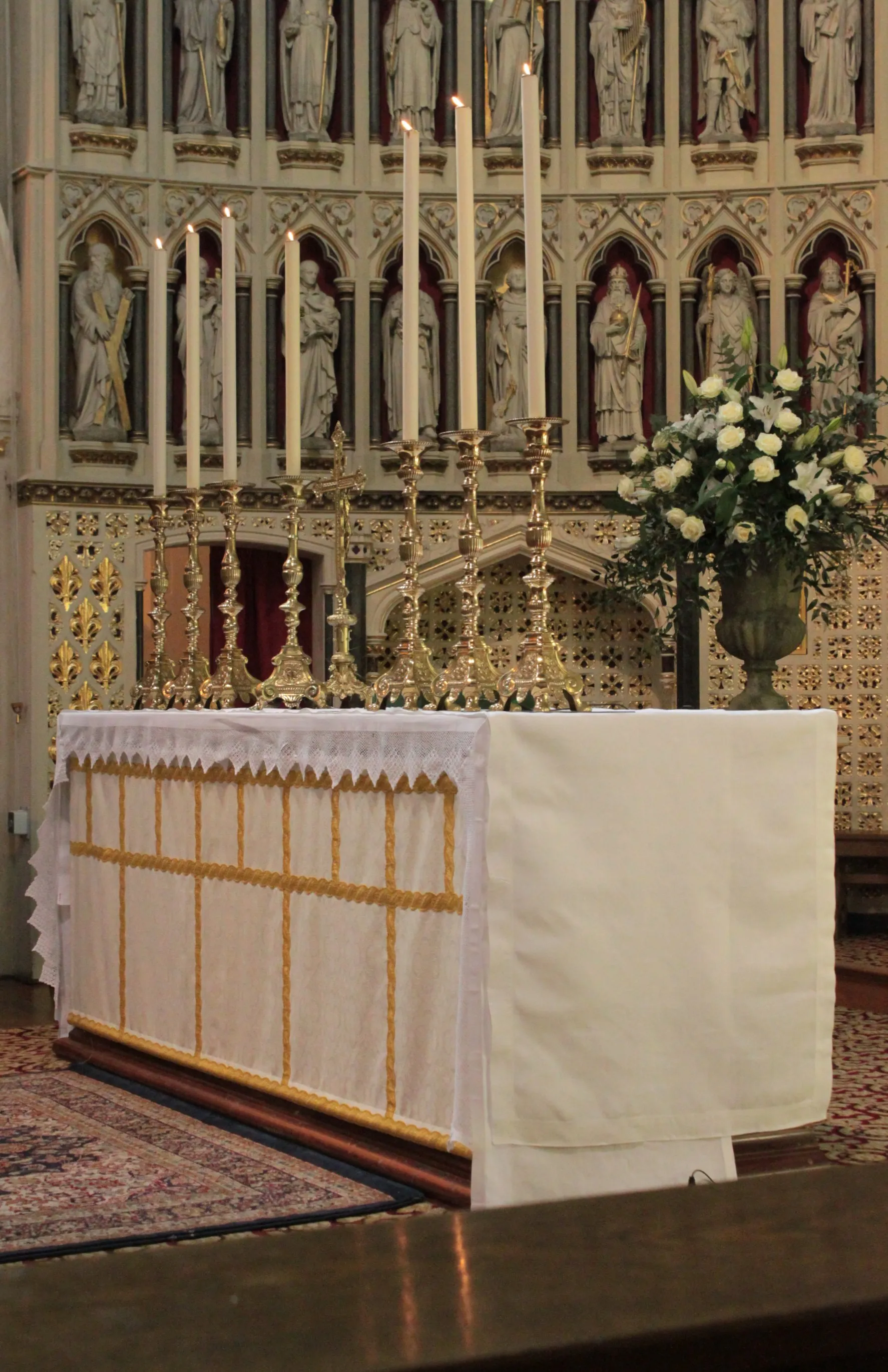 Altar del Oxford Oratory con manteles blancos y dorados, velas altas en candeleros de latón, flores blancas y estatuas neogóticas en las hornacinas traseras.