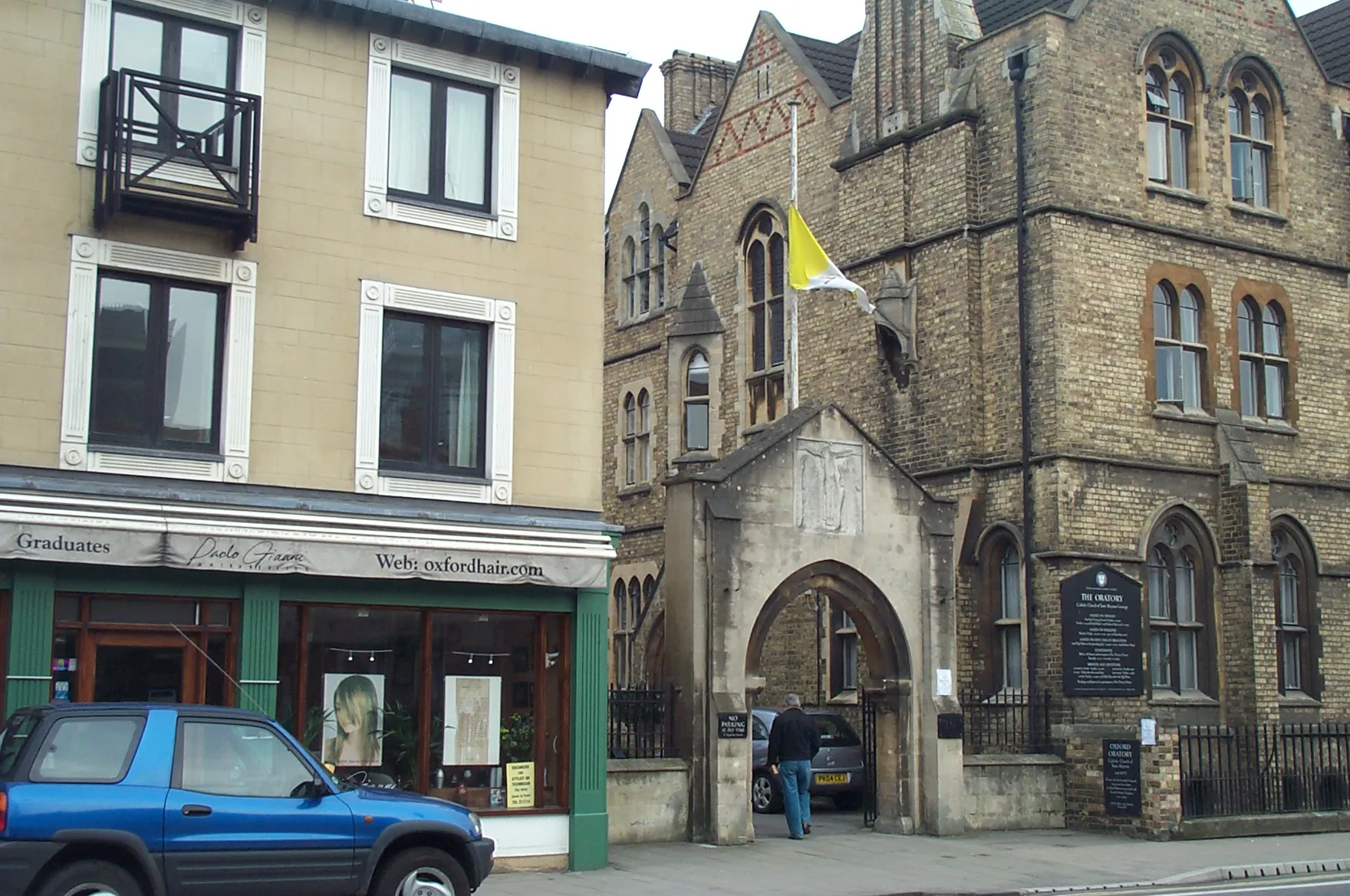 Edificio de iglesia neogótica de piedra con entrada de arco y una bandera del Vaticano amarilla y blanca ondeando arriba, junto a un escaparate con un coche azul aparcado delante.
