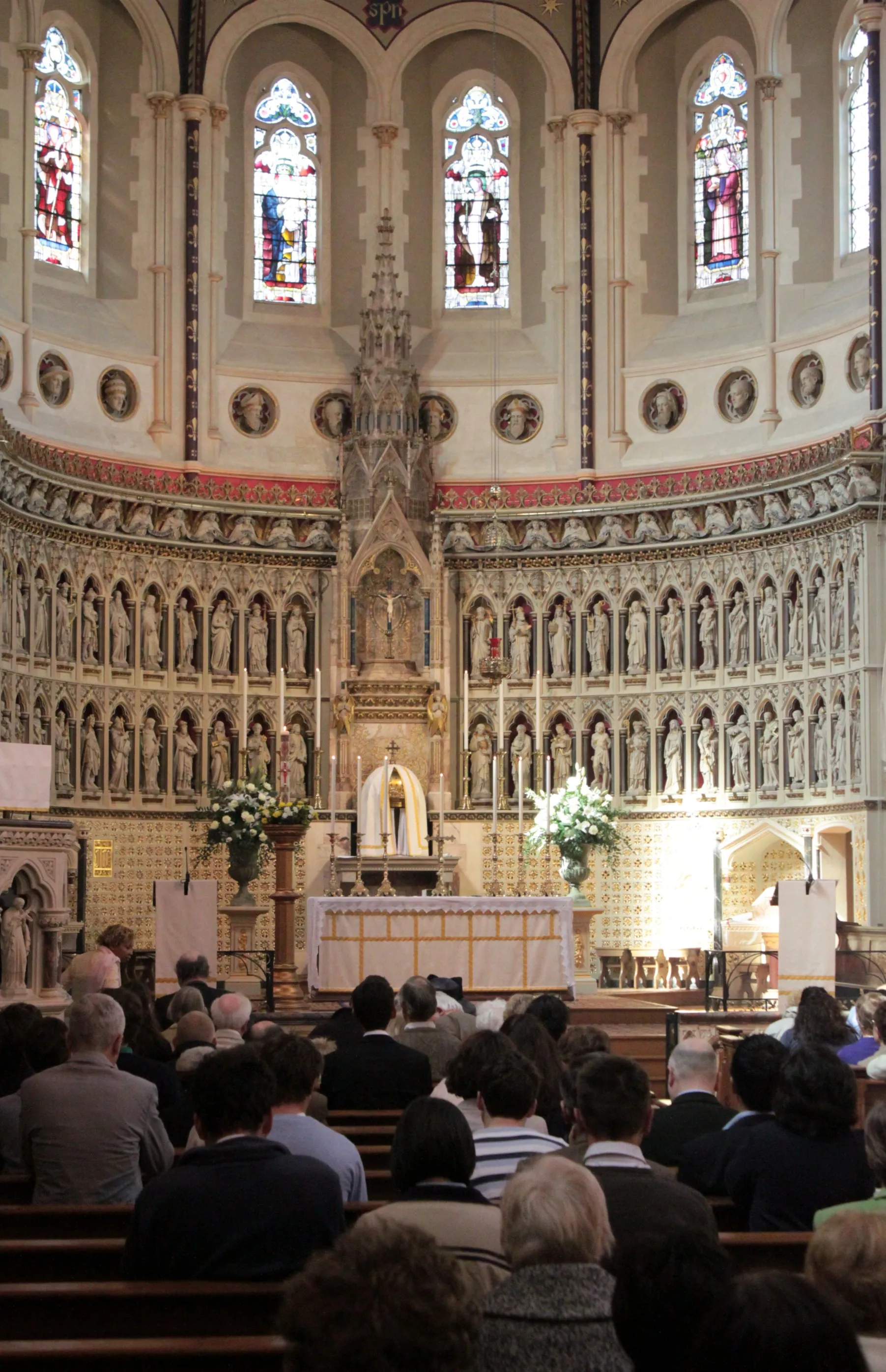 Interior del Oxford Oratory que muestra el altar mayor, cantería neogótica con estatuas en nichos, vidrieras y una congregación sentada en los bancos.