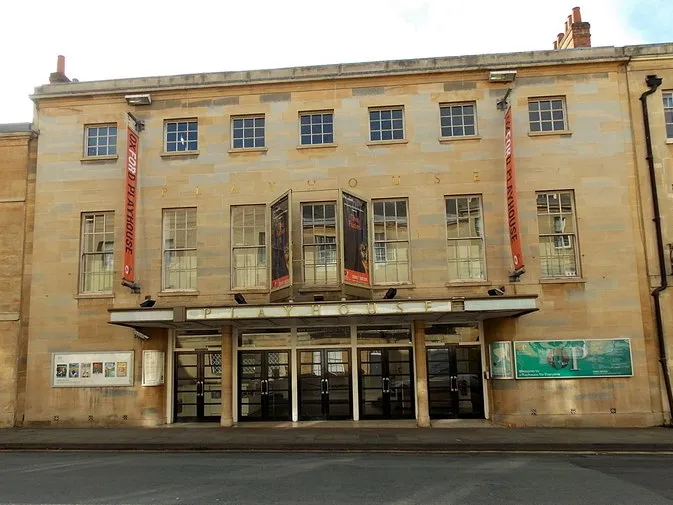Fachada de piedra del teatro Oxford Playhouse con grandes ventanas de guillotina, marquesina de entrada con puertas de cristal y estandartes verticales rojos