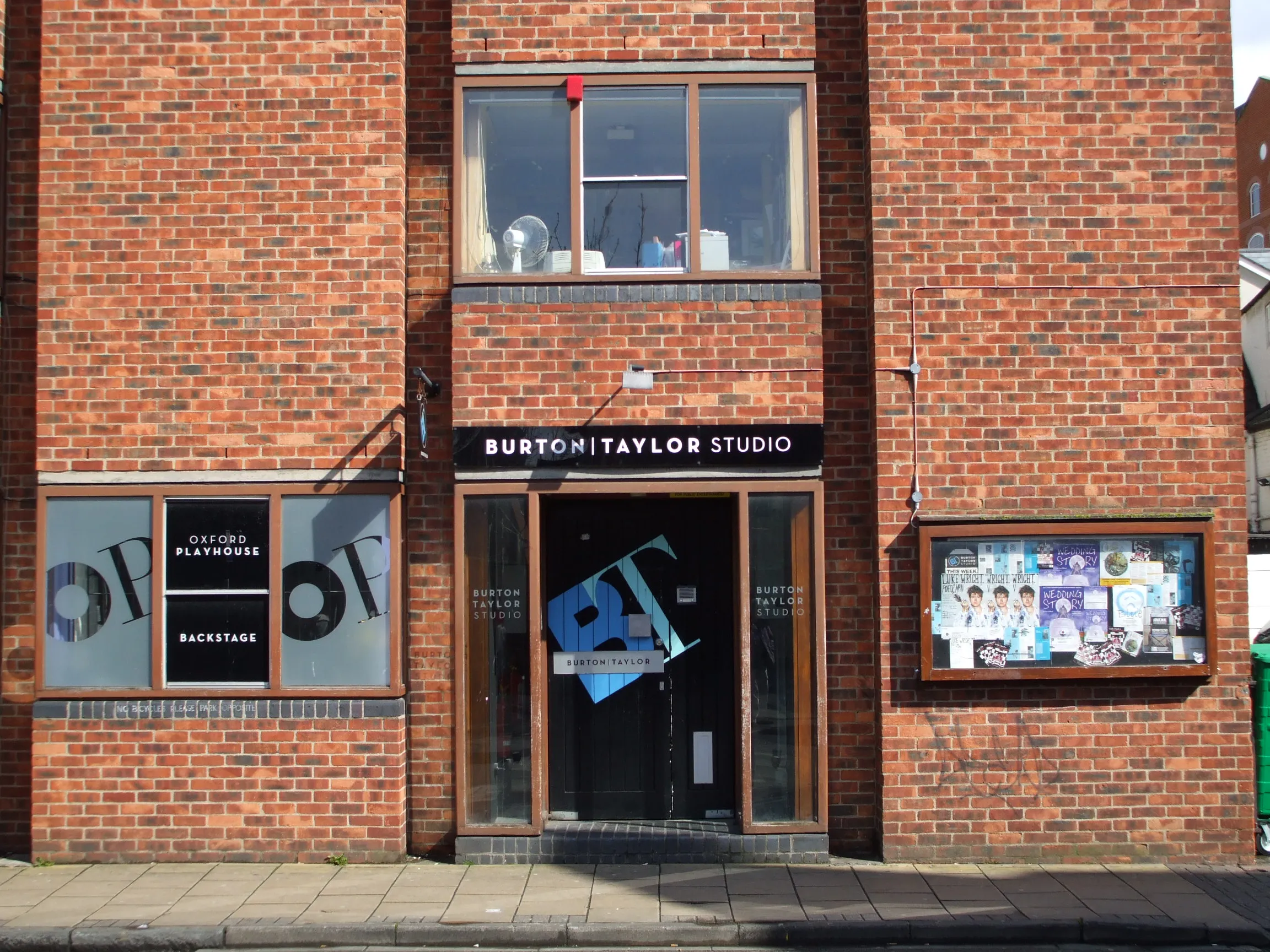 Fachada de ladrillo rojo de la entrada del Burton Taylor Studio con el letrero de recepción del Oxford Playhouse visible en la ventana y un tablón de anuncios a la derecha