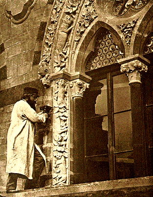 Un hombre con abrigo largo y sombrero está de pie junto a una columna de piedra neogótica profusamente tallada y una puerta arqueada del Oxford University Museum of Natural History.