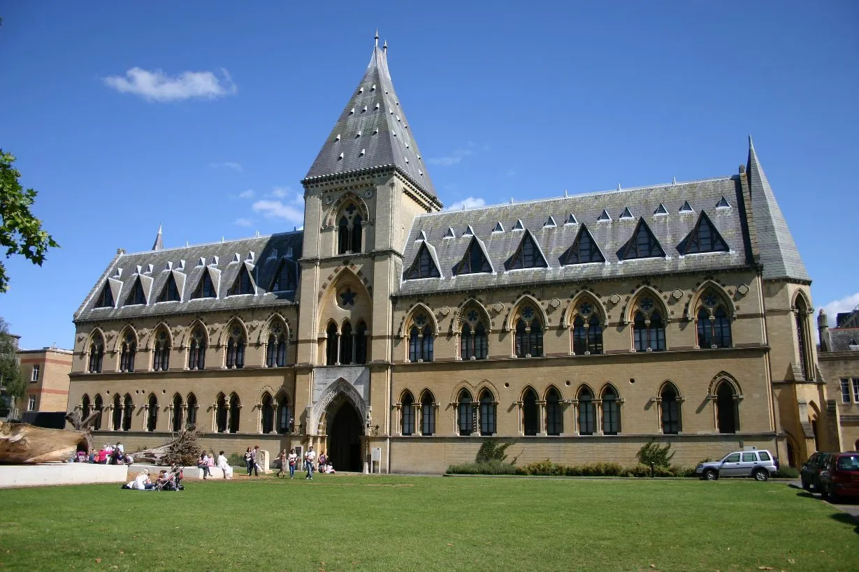 Vista exterior del Oxford University Museum of Natural History, un gran edificio neogótico con una torre central y ventanas arqueadas, frente a un césped verde.