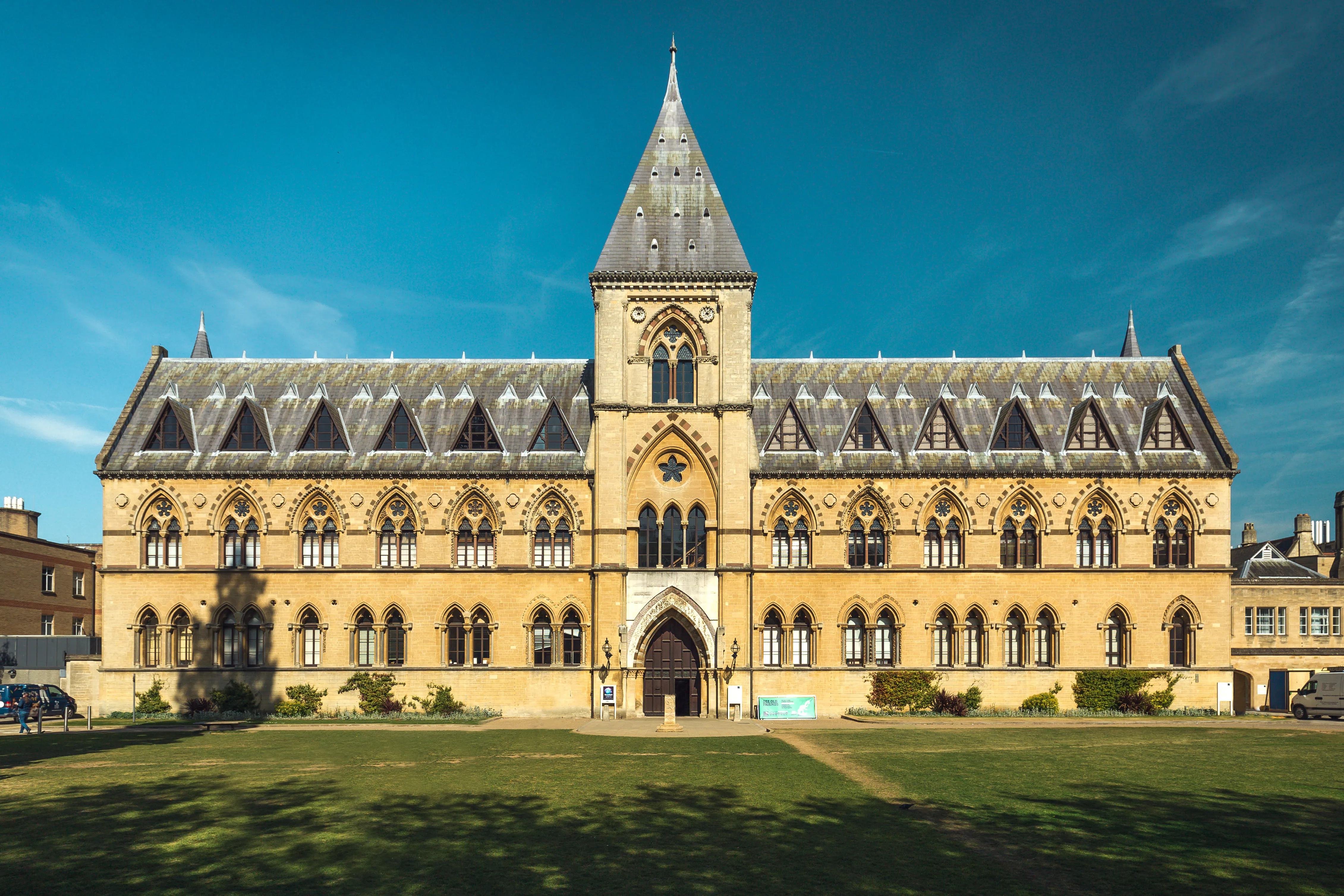 La fachada neogótica del Oxford University Museum of Natural History con una aguja de piedra central, ventanas arqueadas y un césped verde en frente bajo un cielo azul.