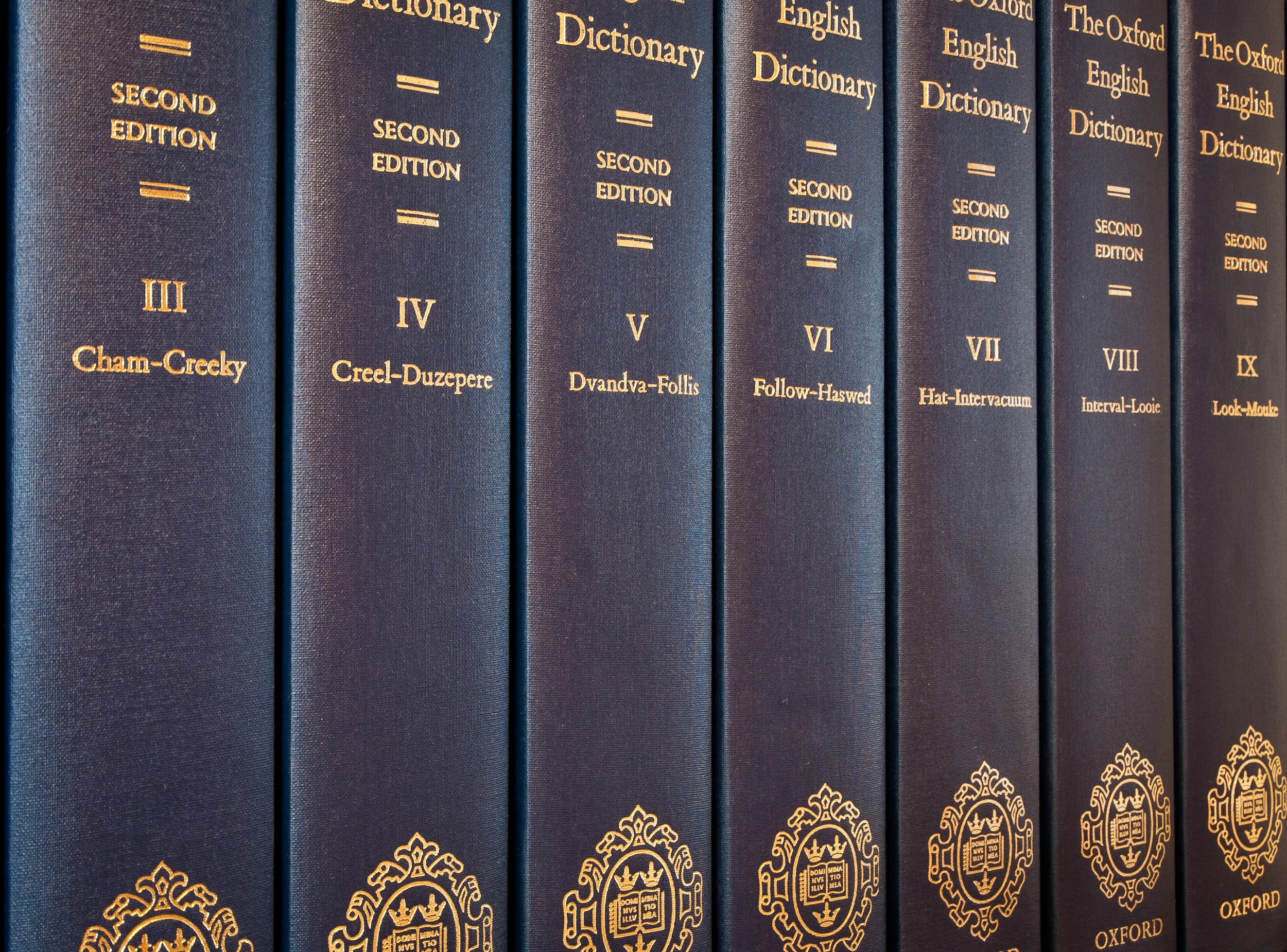 Rząd ciemnoniebieskich tomów w twardej oprawie drugiego wydania Oxford English Dictionary, tomy od III do IX, ze złotymi napisami i ozdobnymi złotymi emblematami na grzbietach