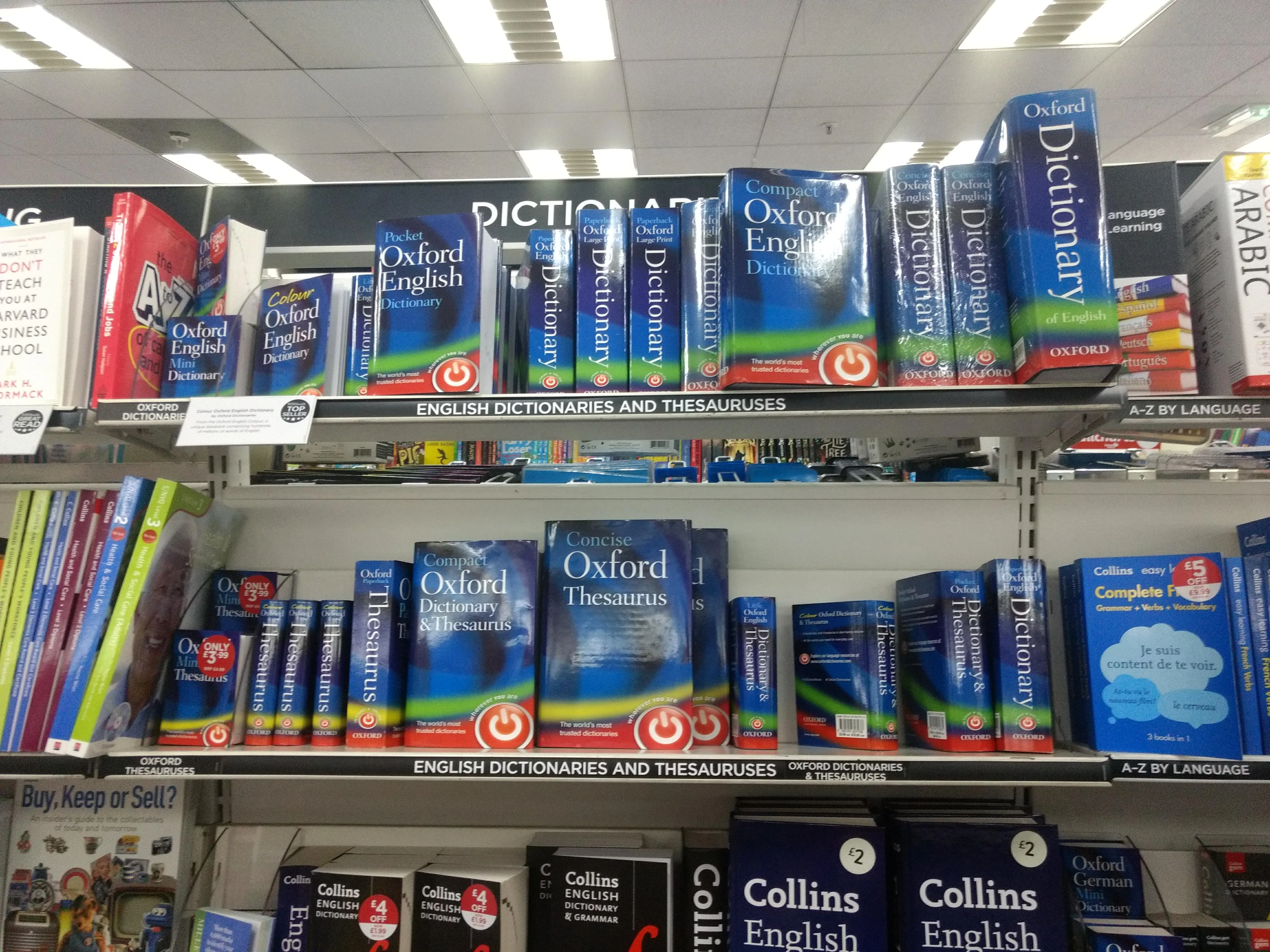 Półki księgarni wypełnione słownikami Oxford English Dictionary i tezaurusami w niebieskich okładkach, z widocznymi również słownikami Collinsa, pod szyldem sekcji 'DICTIONARIES'.