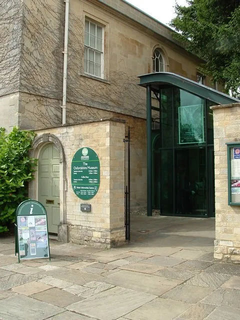 Stone building exterior of The Oxfordshire Museum with a modern glass entrance canopy, a green oval sign reading The Oxfordshire Museum, an information board, and a paved stone courtyard.