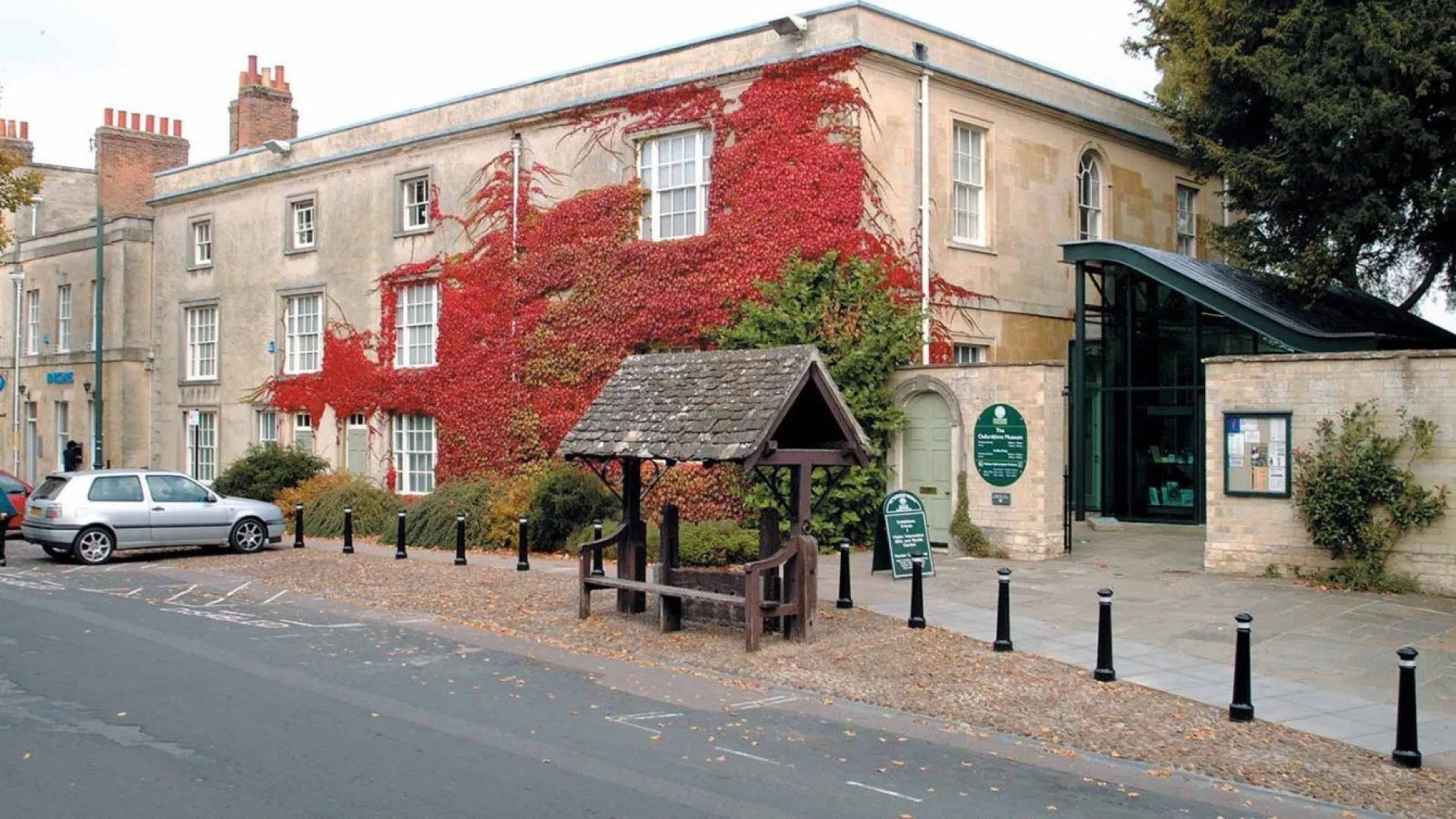 Stone building facade covered in bright red ivy with a small wooden shelter in the foreground and a modern glass entrance to the right.