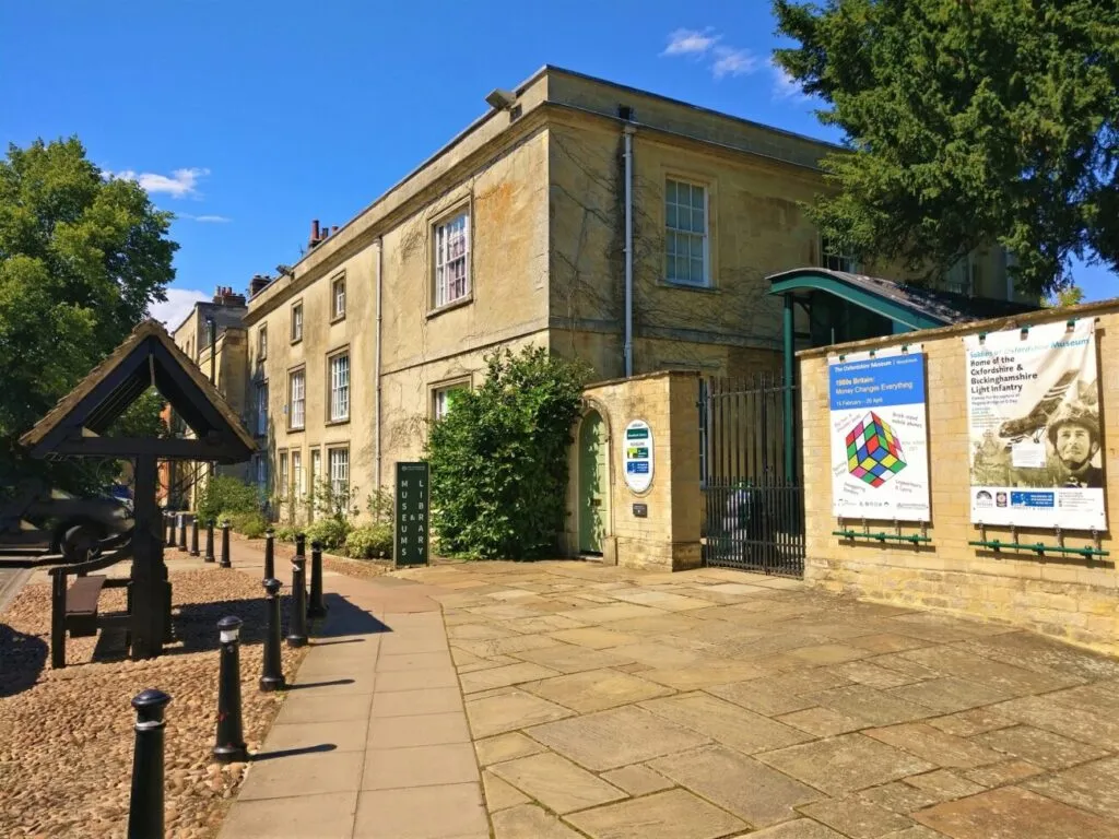 Stone facade of The Oxfordshire Museum with paved courtyard, wooden shelter structure, and exhibition banners on a sunny day