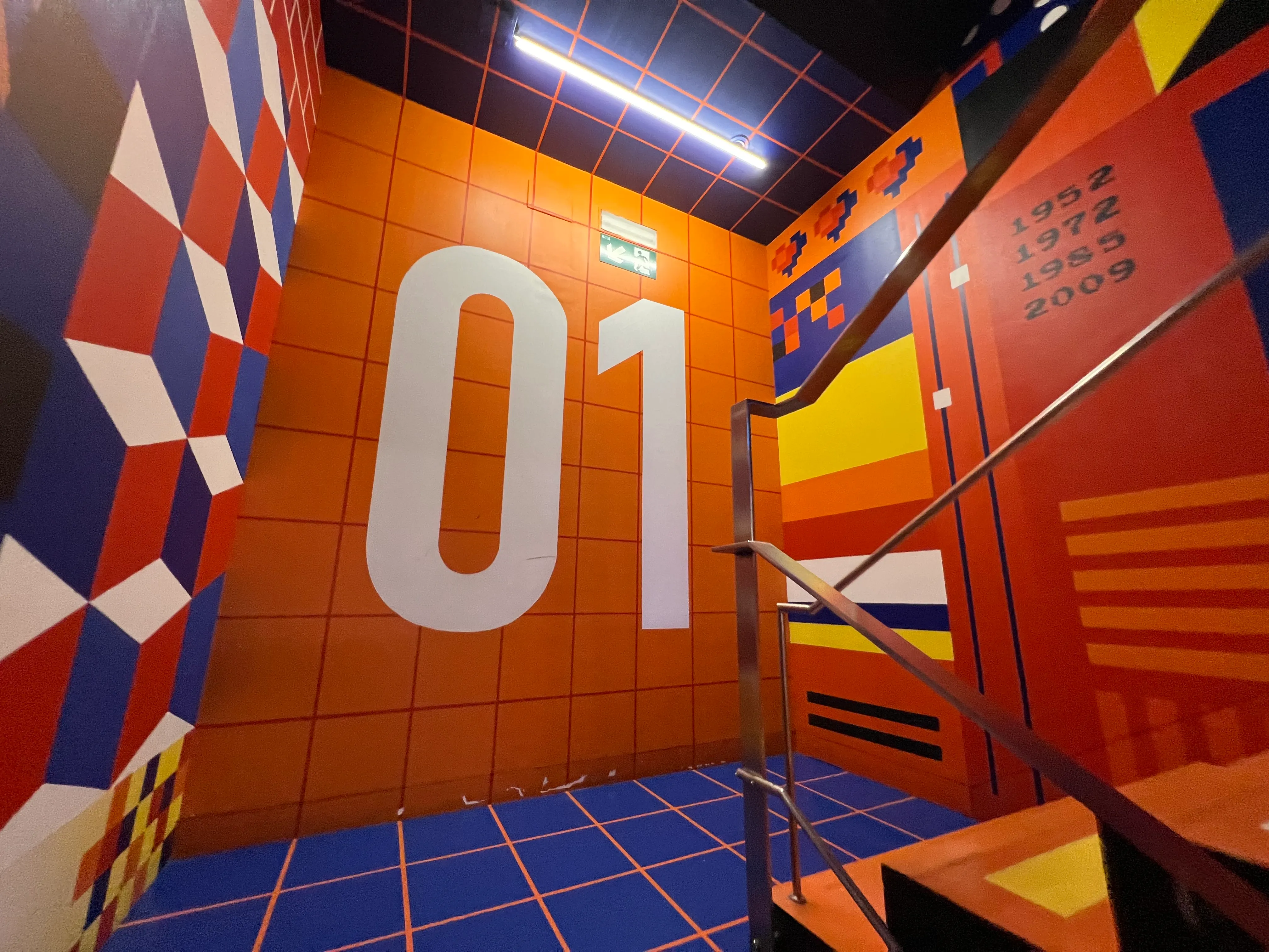 Interior colorido de un museo con un gran "01" blanco en una pared naranja, patrones geométricos de cubos en rojo, blanco y azul, suelo de baldosas azules con líneas de cuadrícula naranjas y una escalera con barandillas de metal.