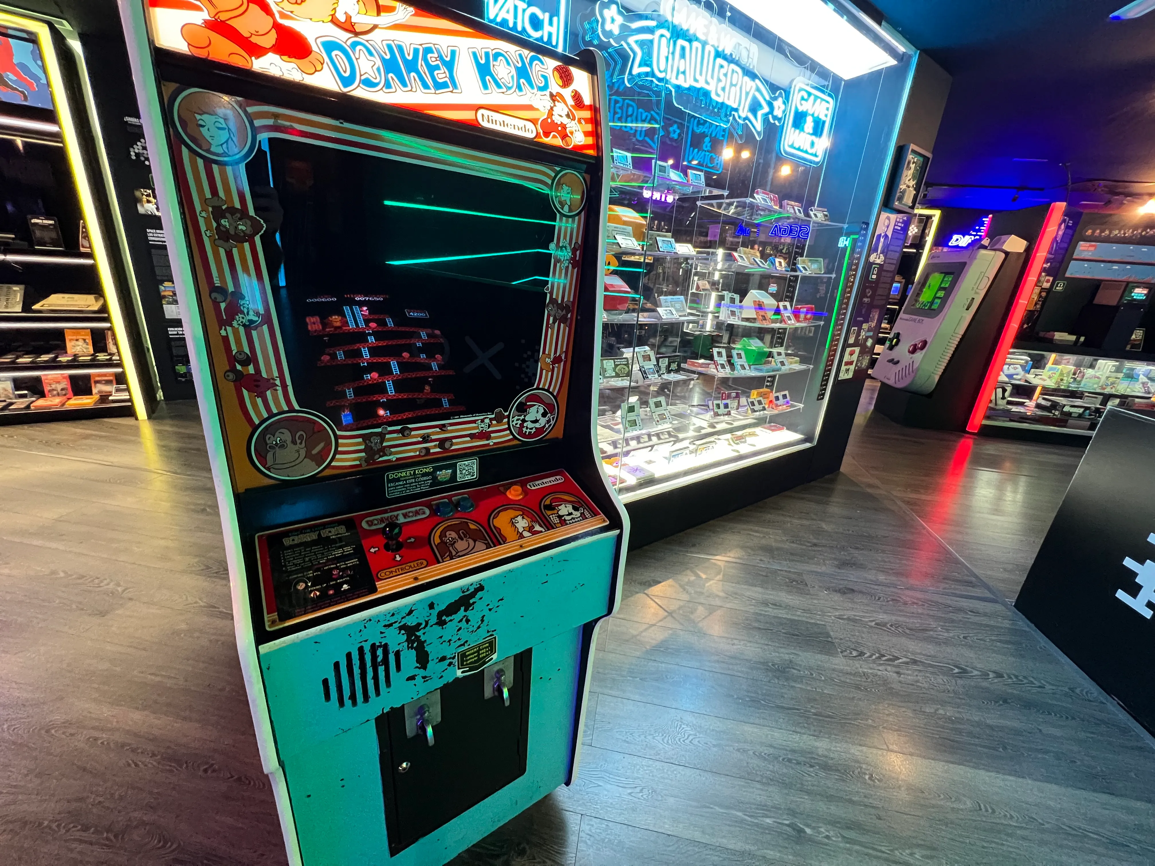 Máquina recreativa de Donkey Kong expuesta en un museo de videojuegos con una vitrina de cristal con objetos de colección y luces de neón al fondo