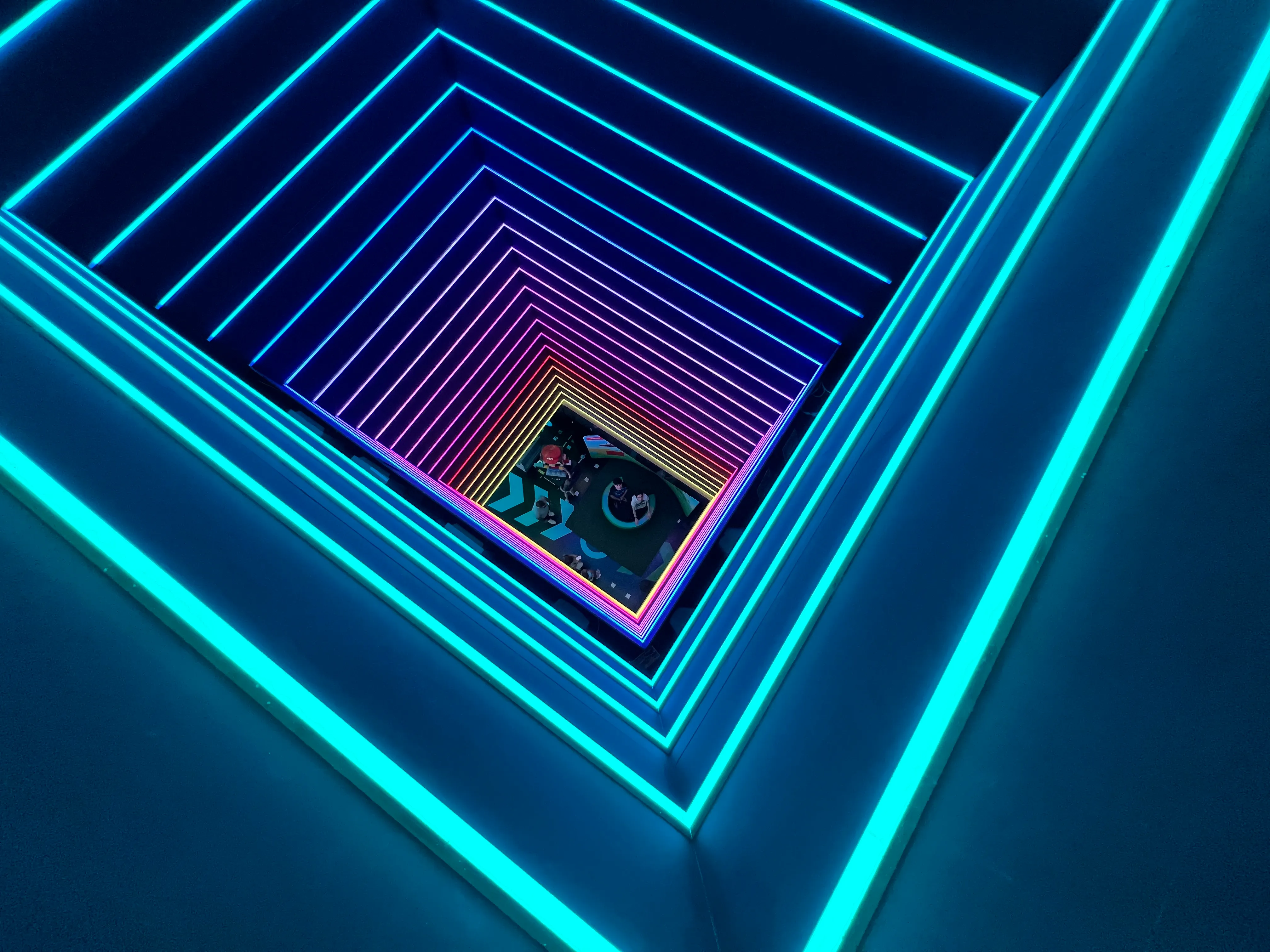 Pasillo con túnel iluminado por neón con marcos rectangulares concéntricos de luces LED cian y azules que crean un efecto de perspectiva infinita, con una pantalla de juego retro visible al fondo.