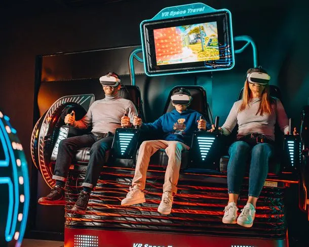 Three people wearing VR headsets sitting in a gaming simulator with a screen showing a video game