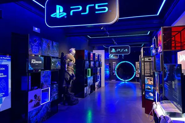Interior hallway of a video game museum with blue neon lighting, PS5 and PS4 signs, arcade machines, and a life-sized character statue