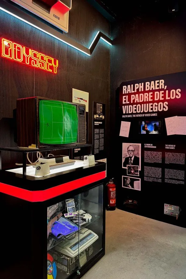 A vintage Odyssey video game console with a green screen on display under a neon 'ODYSSEY' sign, next to a wall with text about Ralph Baer, the father of video games.