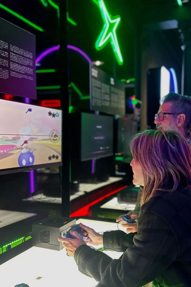Two people playing a video game on a console with a screen showing a colorful game, in a room with neon lights and arcade machines.