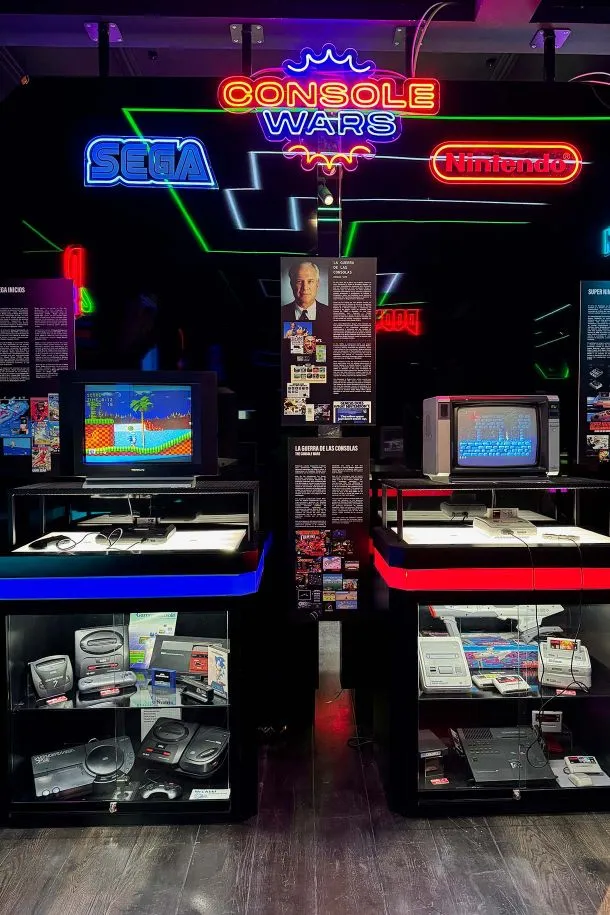 Neon sign reading 'Console Wars' with Sega and Nintendo logos above vintage gaming consoles on display