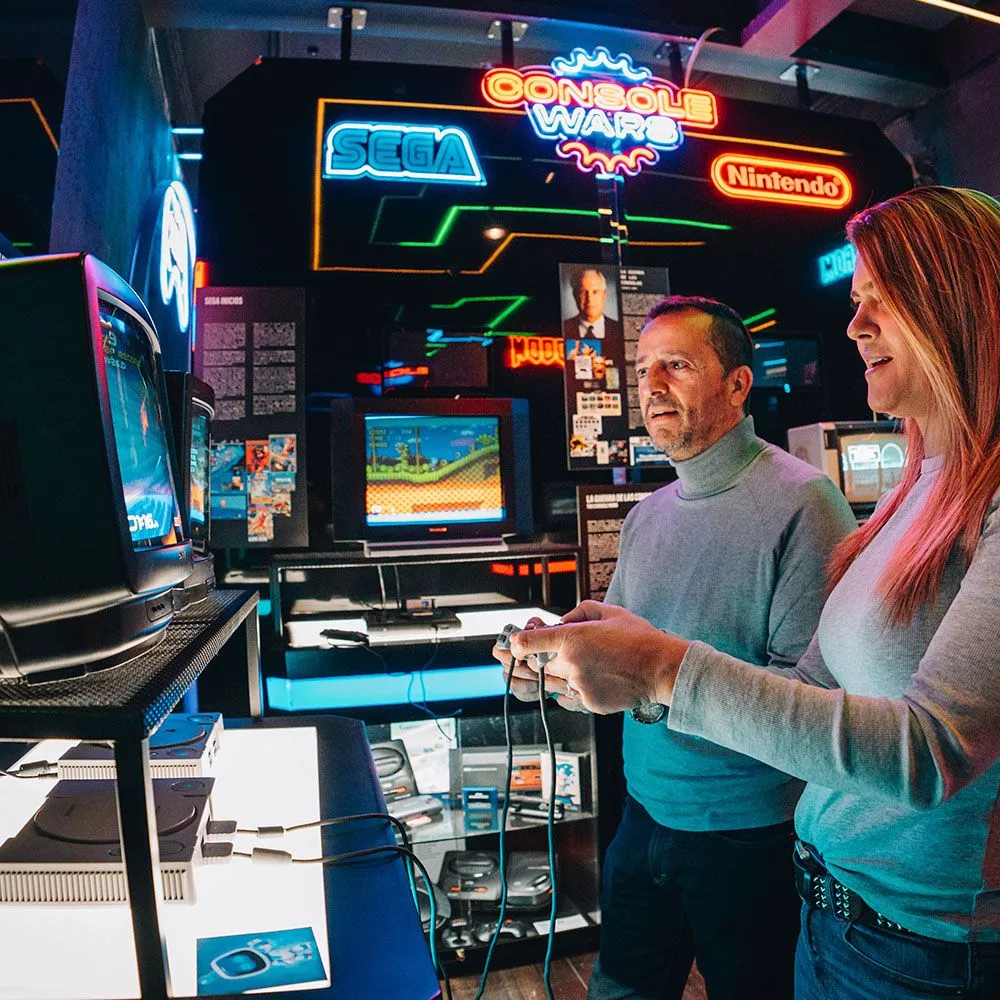 Twee mensen die een vintage arcadespel spelen in een museum met neonreclames voor Sega, Nintendo en Conquest War