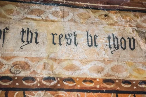 Zbliżenie na średniowieczne malowidło ścienne z tekstem w piśmie blackletter o treści 'of thi rest be thou', obramowane dekoracyjnymi malowanymi krawędziami na postarzanym tynku.