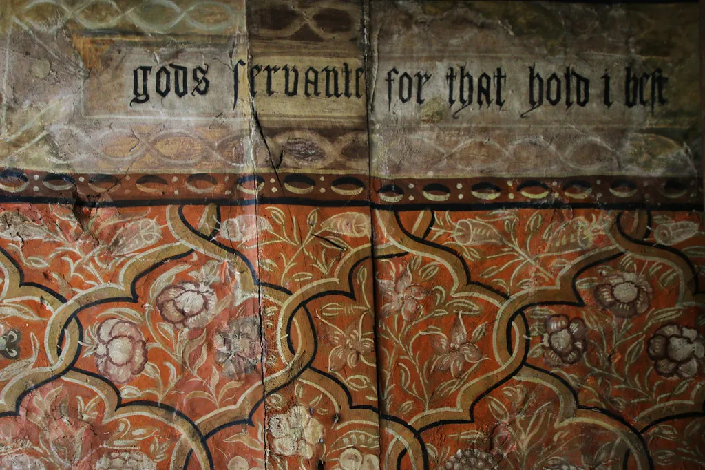 Zbliżenie na elżbietańskie malowidła ścienne z gotyckim tekstem typu blackletter o treści 'gods servants for that hold' nad zdobnymi czerwono-kremowymi motywami roślinnymi na postarzałym tynku