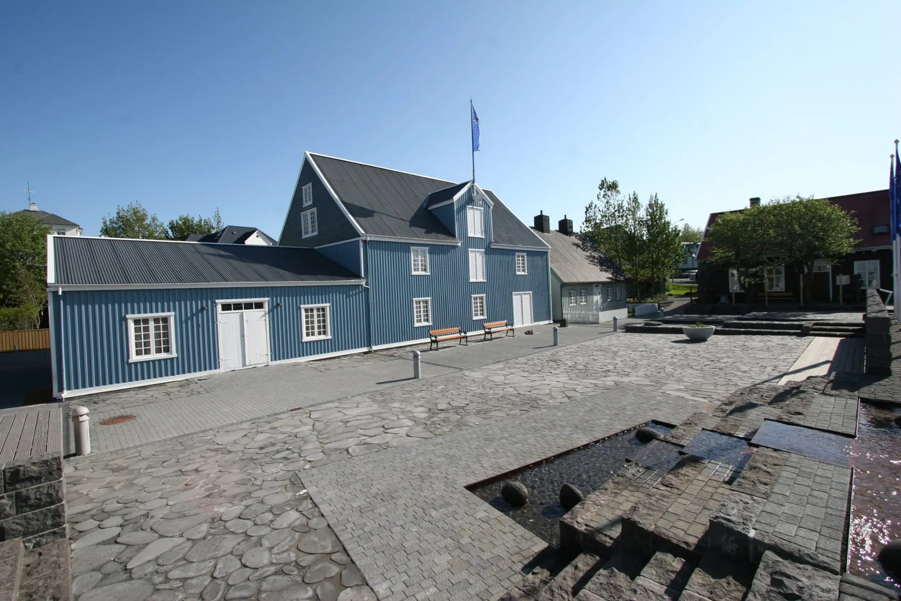 A blue wooden museum building with white trim and a stone-paved courtyard featuring archaeological ruins in the foreground.